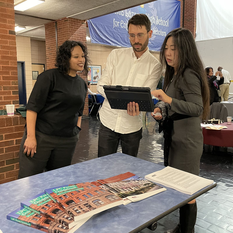 Three people stand looking at content on a tablet device